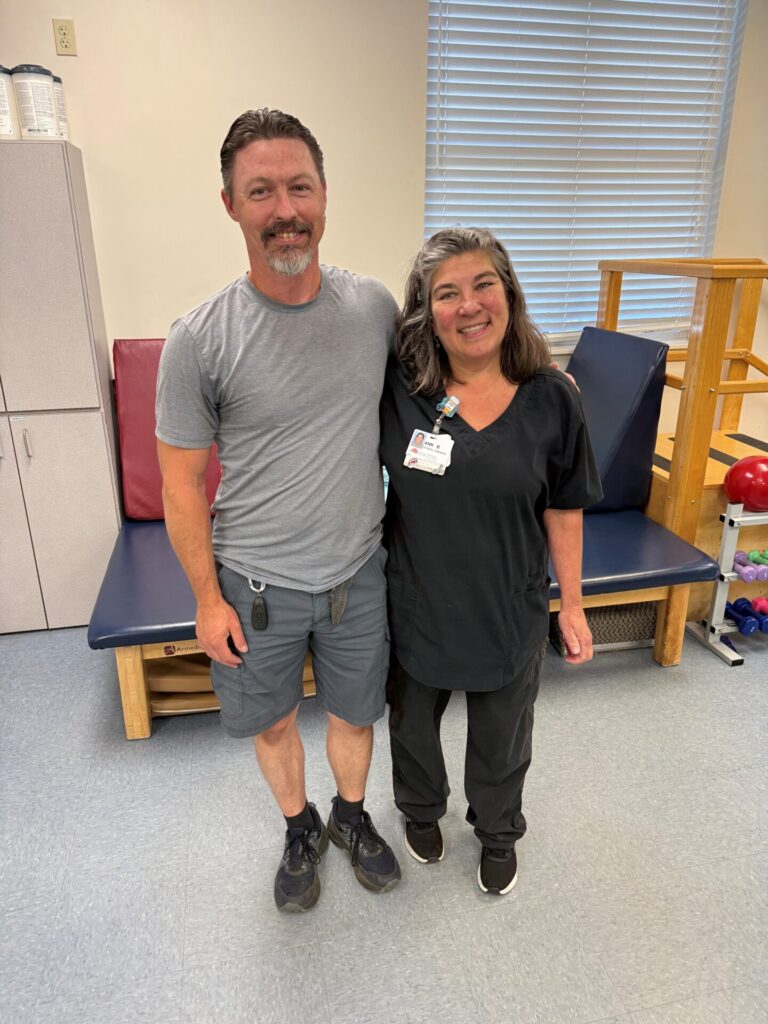 A man in a gray t-shirt and shorts stands smiling next to a woman in black medical scrubs with a name badge, both inside a bright clinical room with gym equipment in the background.
