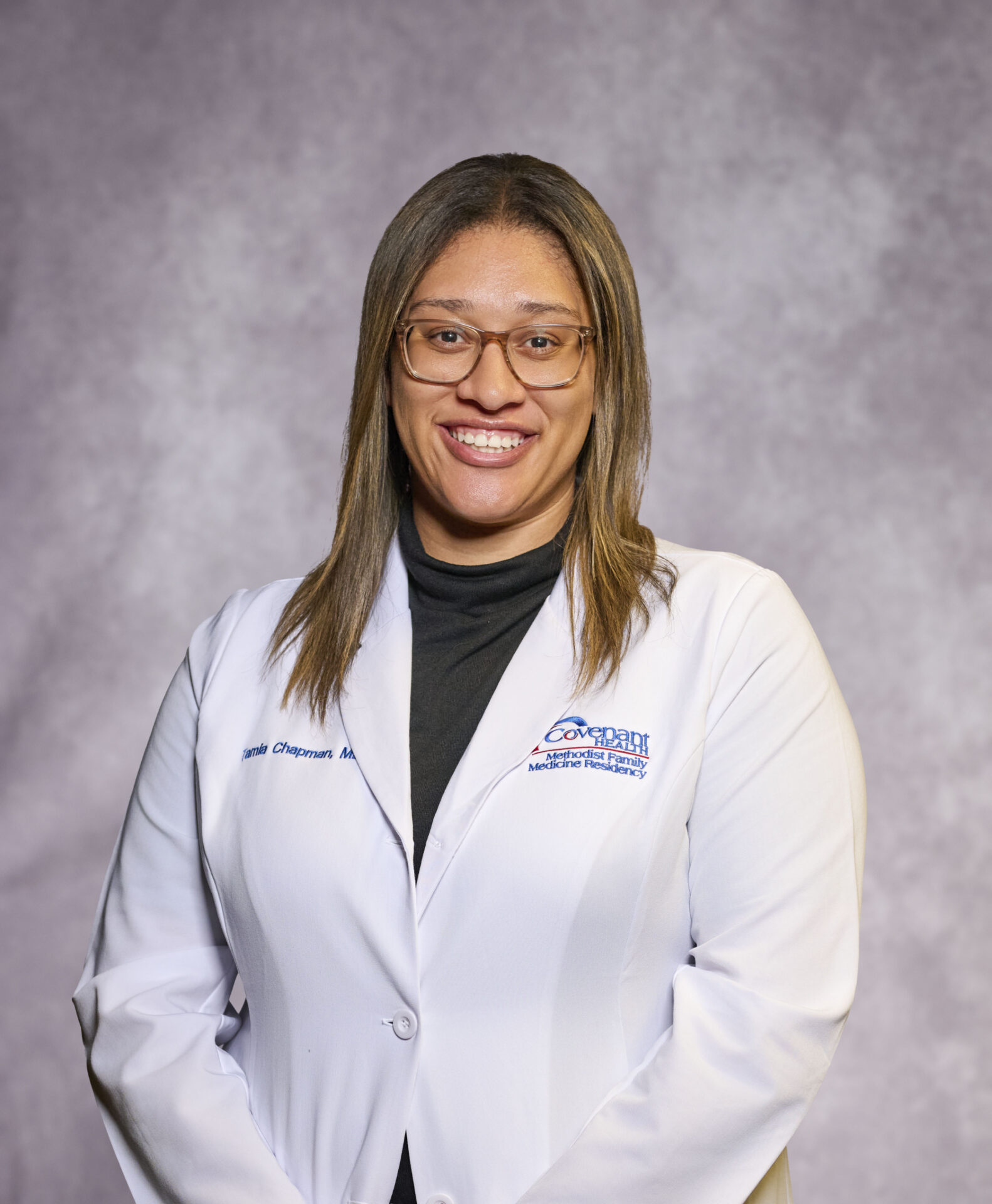 A woman wearing glasses and a white lab coat stands smiling in front of a gray, textured backdrop.