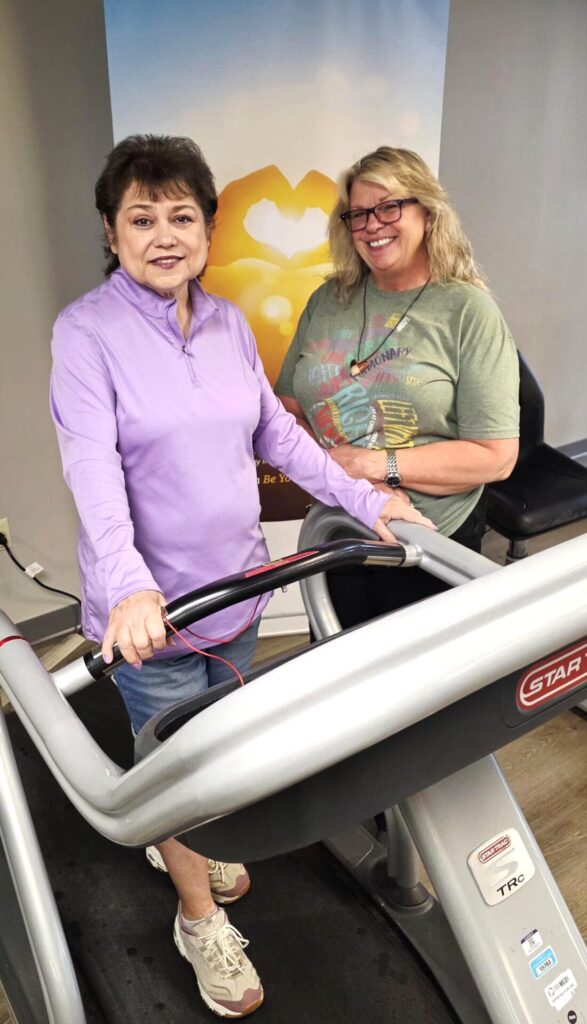 Two women smile and stand next to each other at a treadmill in a gym, with one woman holding the treadmill handles. A bright, heart-shaped poster is visible on the wall behind them.