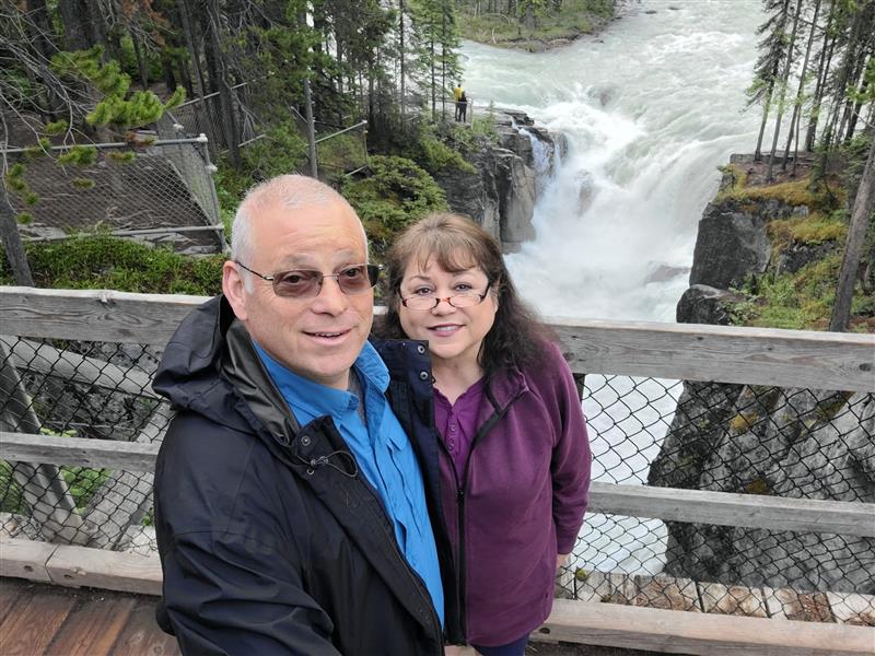A man and woman smile on a wooden bridge overlooking a waterfall surrounded by trees and rocks.