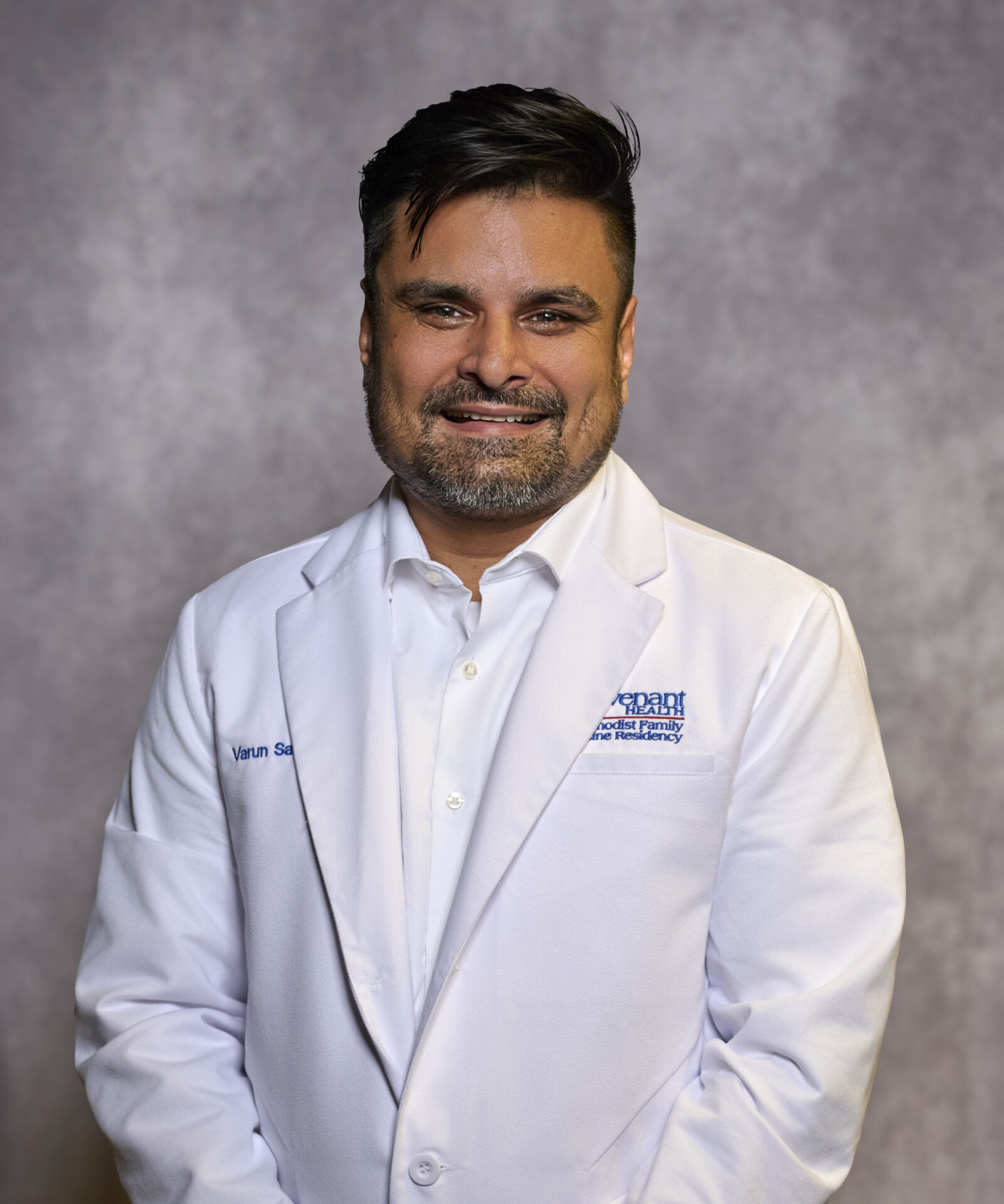 A man with short dark hair and a beard, wearing a white lab coat and white shirt, stands in front of a neutral gray background, smiling at the camera.