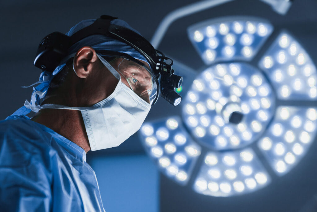 A surgeon wearing a mask, headlamp, and surgical cap focuses intently during surgery, with bright operating room lights illuminating the background.