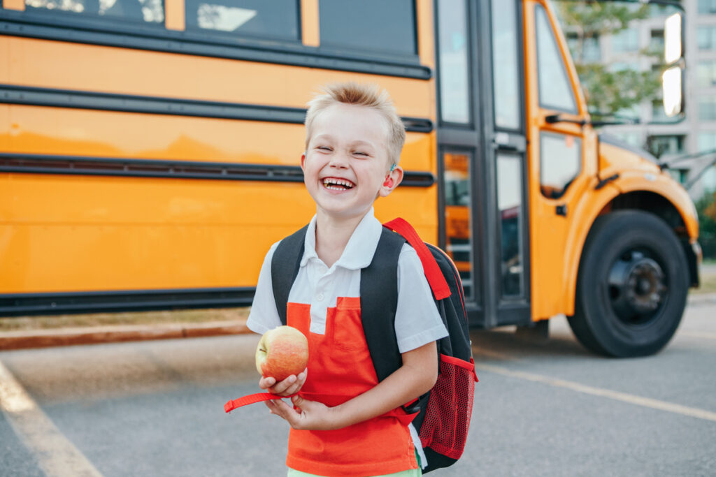 Smiling young boy with a backpack holds an apple while standing in front of a yellow school bus on a sunny day.