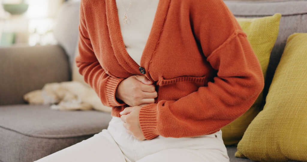 A person wearing an orange cardigan and white pants is sitting on a couch, holding their stomach with both hands as if experiencing pain or discomfort. There are yellow pillows and a dog in the background.