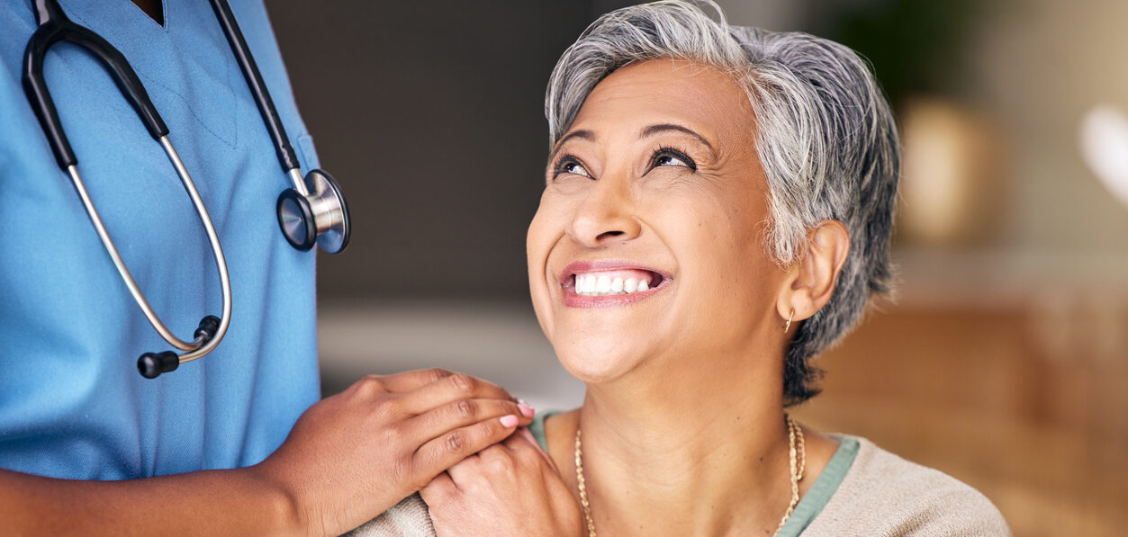 An older woman smiles while holding hands with a nurse in blue scrubs, who stands beside her with a stethoscope around their neck. The scene conveys comfort and support.