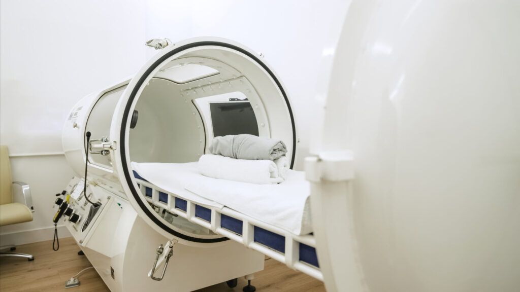 A hyperbaric oxygen chamber with white towels and a folded blanket on the bed inside, in a clean medical room with white walls and a chair in the corner.