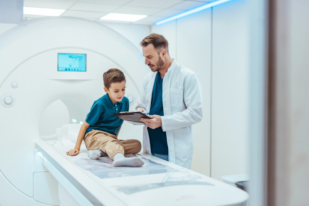 A young boy sits on an MRI scanner bed while a doctor in a white coat shows him information on a clipboard in a medical imaging room.