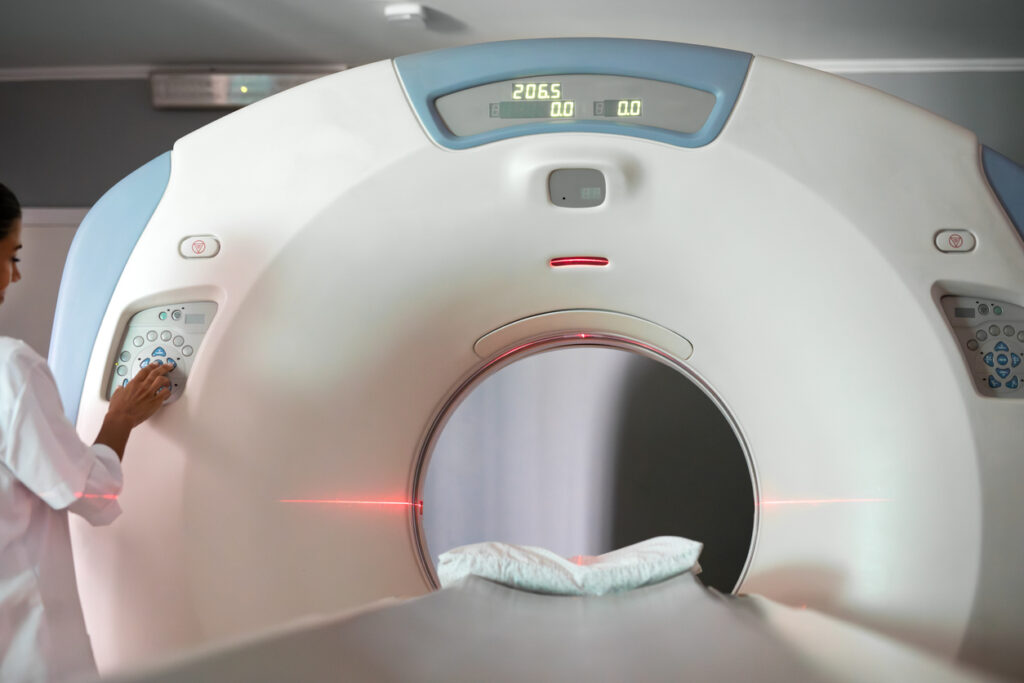 A medical professional operates a large MRI or CT scanner in a hospital room, with a pillow placed on the machine&rsquo;s bed and red laser lines visible inside the scanning tunnel.