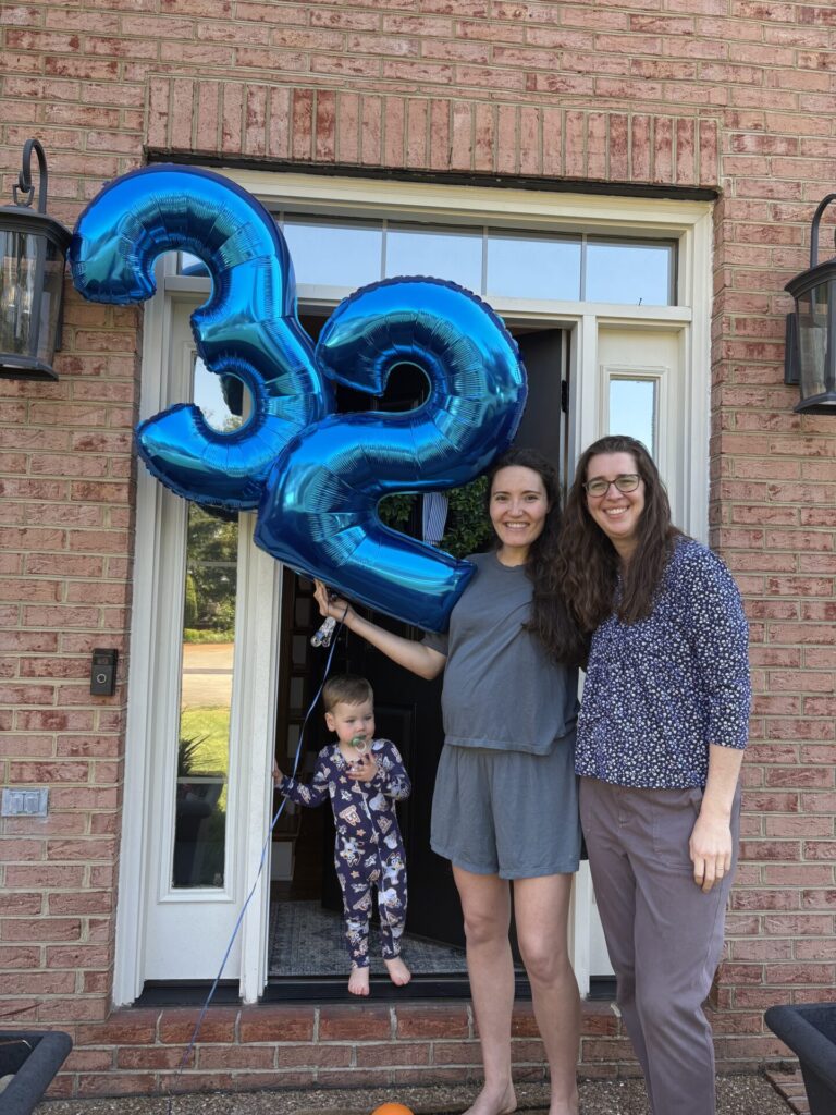 Two women stand in a doorway, smiling, holding large blue 32 balloons. A small child in pajamas stands nearby. They are in front of a brick house with two wall lanterns on either side of the door.