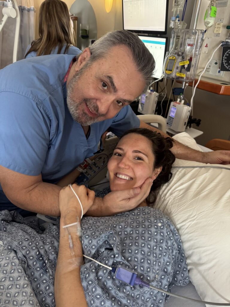 A woman in a hospital bed smiles while a man in scrubs gently holds her face, celebrating a moment of therapy recovery. Medical equipment and monitors are visible in the background.