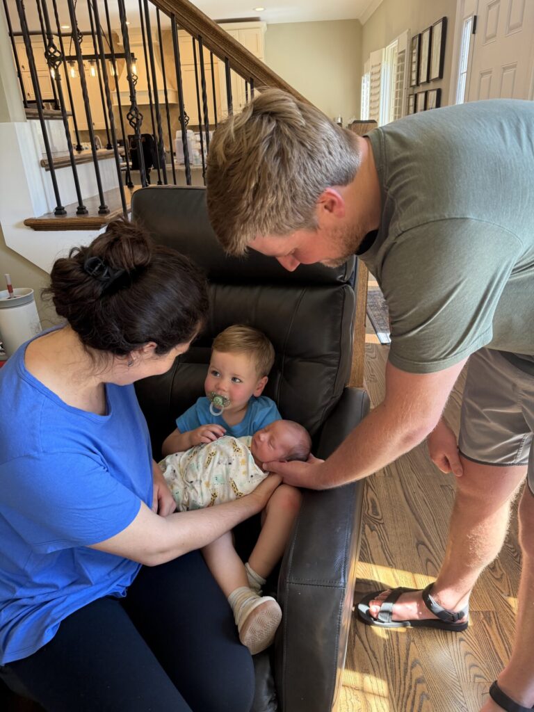 A woman sits in a chair holding a baby, while a toddler with a pacifier sits beside her. A man stands nearby, gently touching the baby’s head as if offering comfort and support during therapy recovery. They are in a sunlit living room.