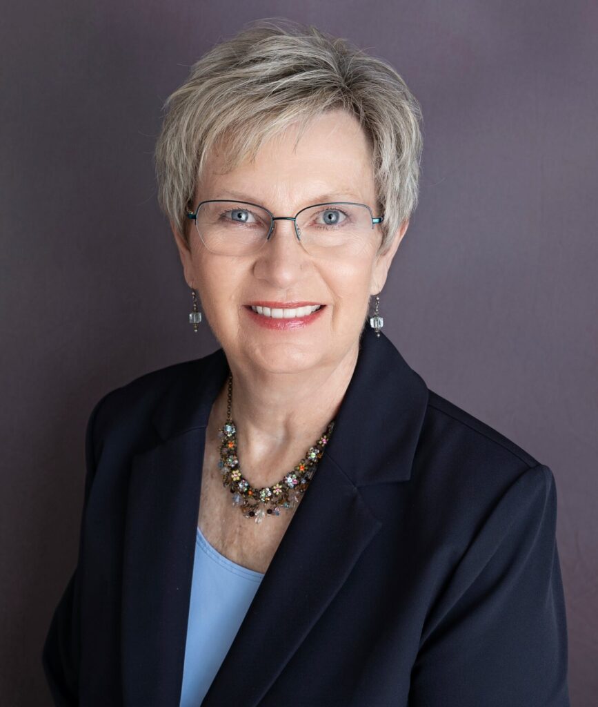 Smiling woman with short gray hair, glasses, and a dark blazer over a light blue top, wearing a colorful beaded necklace, posing in front of a plain gray background.