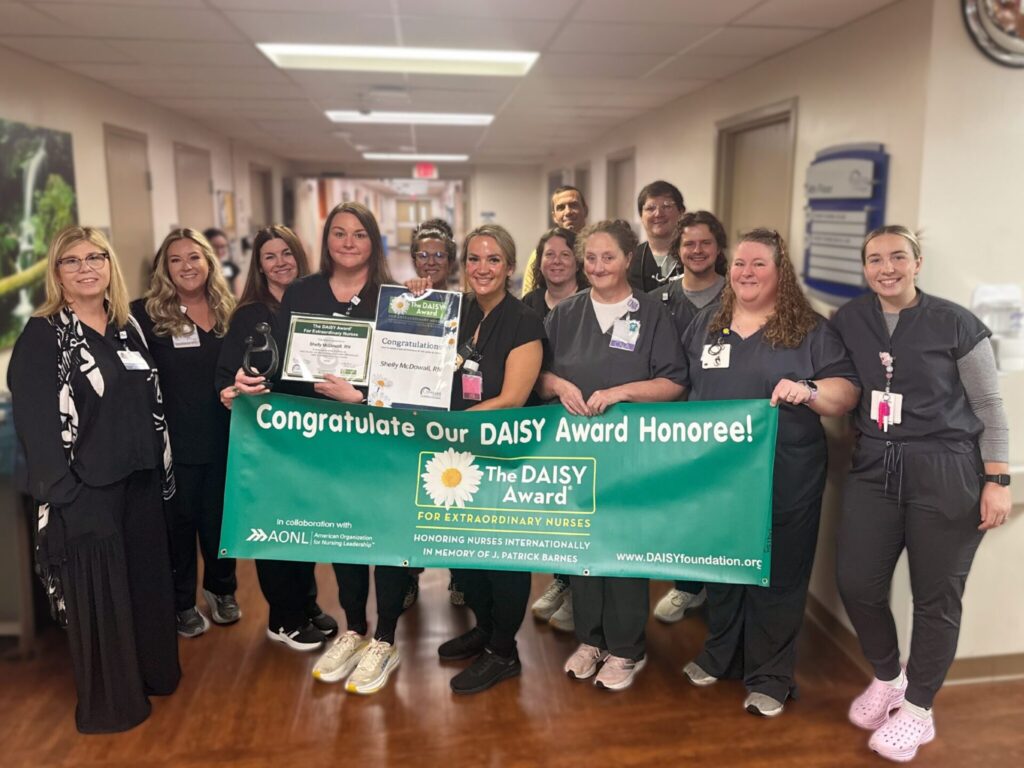 A group of healthcare workers in scrubs and badges stand in a hallway, celebrating DAISY Award Nominee Michelle Martin RN as they hold a green banner that reads, Congratulations Our DAISY Award Honoree! and smile at the camera.