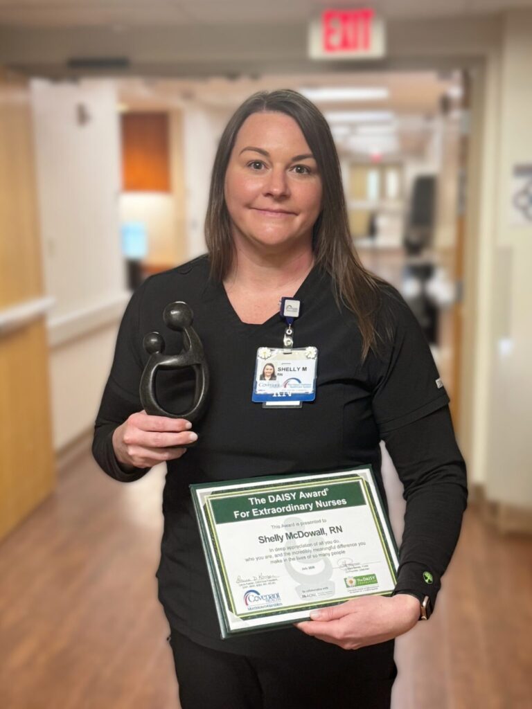 A nurse in black scrubs holds a DAISY Award trophy and certificate in a hospital hallway, smiling at the camera—celebrating DAISY Award Nominee Michelle Martin RN and her outstanding dedication to patient care.