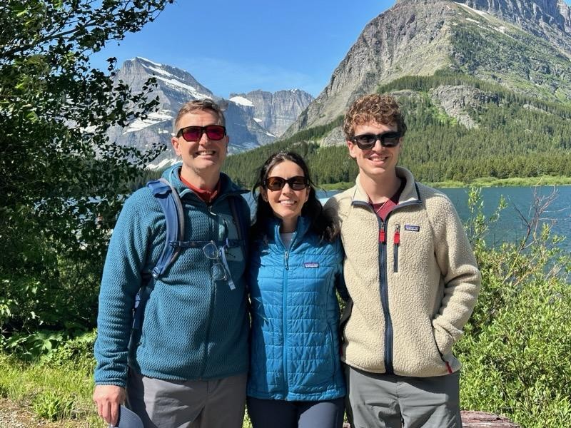 Three people wearing sunglasses and jackets stand outdoors, smiling, with a lake, trees, and snow-capped mountains in the background on a sunny day.