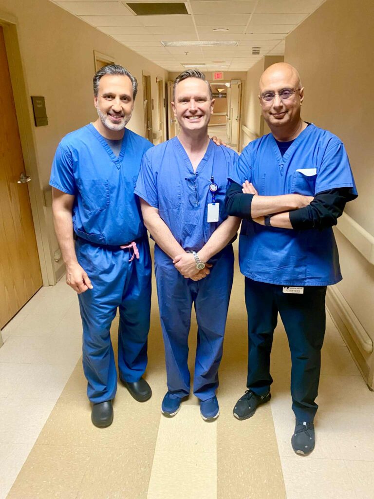 Three people in blue scrubs stand smiling in a hospital hallway with beige walls and floors, posing for a group photo.