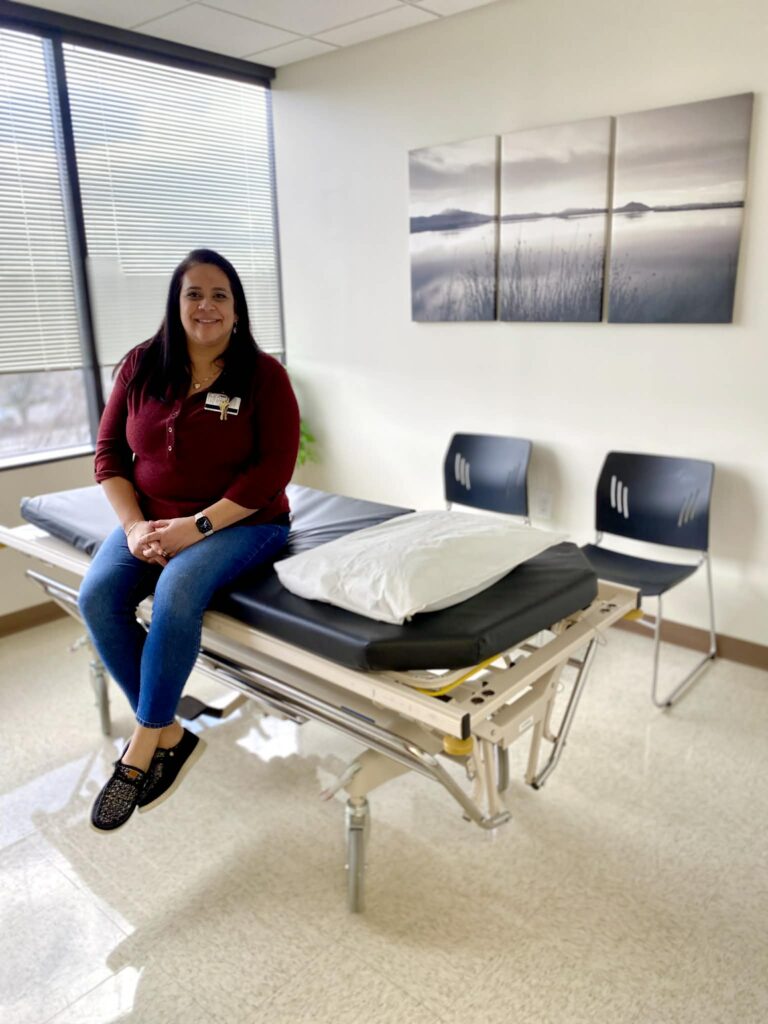 A woman sits on a medical exam table in a bright room with two chairs, a pillow, and landscape art on the wall, waiting to discuss her swimmer&rsquo;s ear with the healthcare provider.