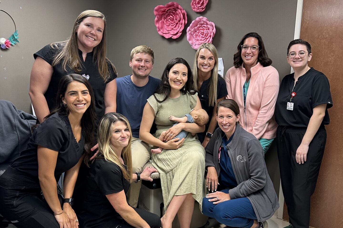 A group of nine adults, mostly women, and one baby pose and smile together in a medical office decorated with large pink paper flowers on the wall.