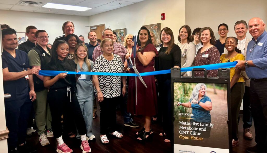 A diverse group of people smiles as they cut a blue ribbon at a clinic open house event. A sign in front promotes the Methodist Family Metabolic and OMT Clinic Open House, highlighting services for conditions such as swimmer’s ear.