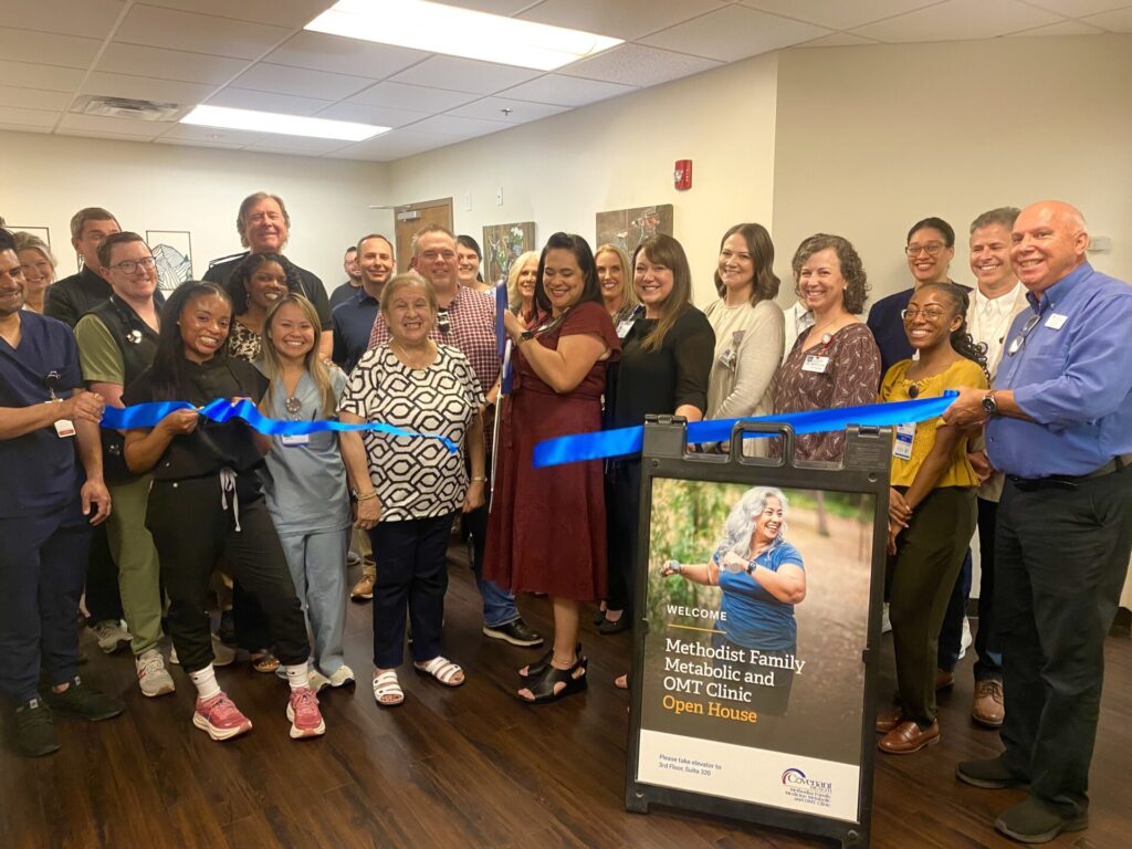 A diverse group of people smiles as they cut a blue ribbon at a clinic open house event. A sign in front promotes the Methodist Family Metabolic and OMT Clinic Open House, highlighting services for conditions such as swimmer&rsquo;s ear.