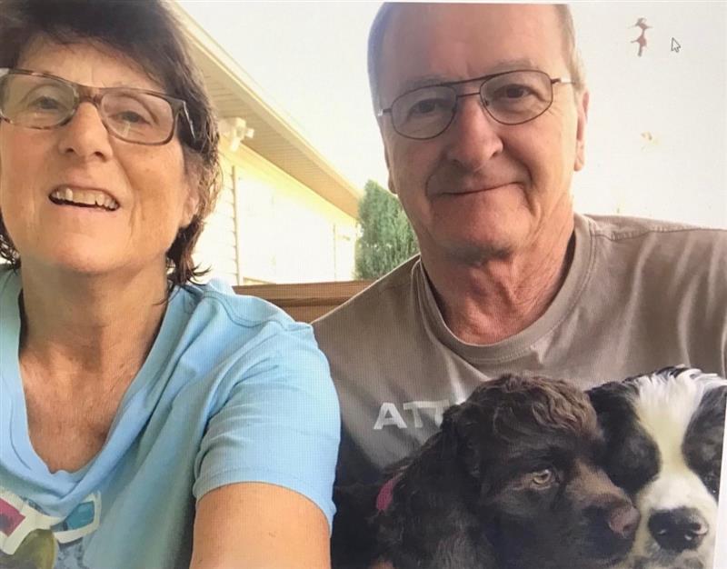 A smiling older woman and man wearing glasses sit close together outside, each holding a puppy in their laps. The background shows part of a house and trees.