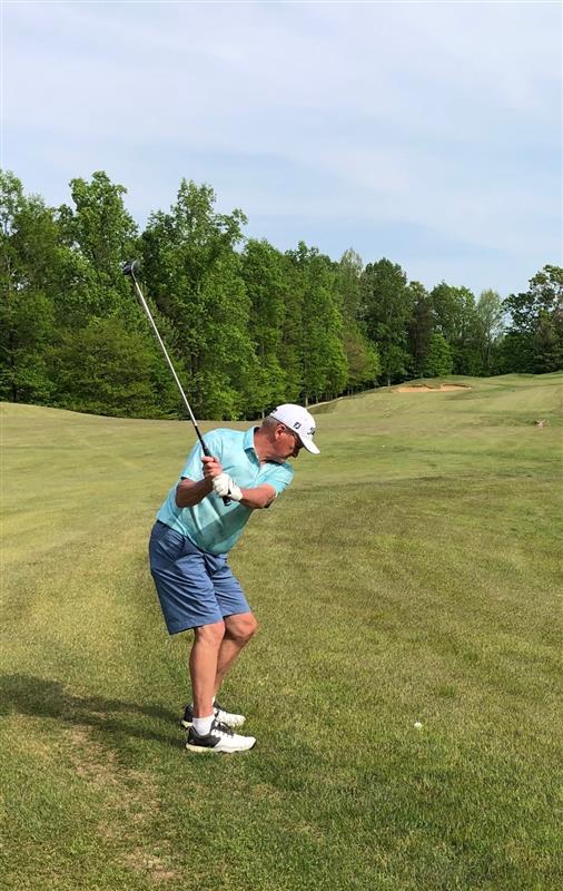 Man in a light blue shirt, shorts, and cap playing golf on a grassy course, preparing to swing his club with trees and an open fairway in the background.