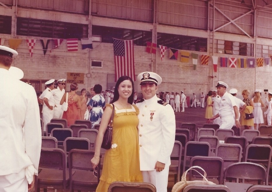 A woman in a yellow dress stands next to a man in a white naval uniform inside a large hall with rows of chairs and people in similar uniforms; flags hang above them.