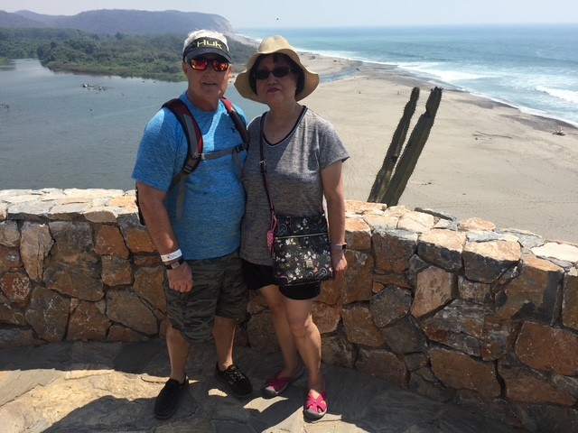 A man and woman stand together by a stone wall overlooking a beach and ocean on a sunny day. Both wear hats and sunglasses, with the woman holding a floral purse.