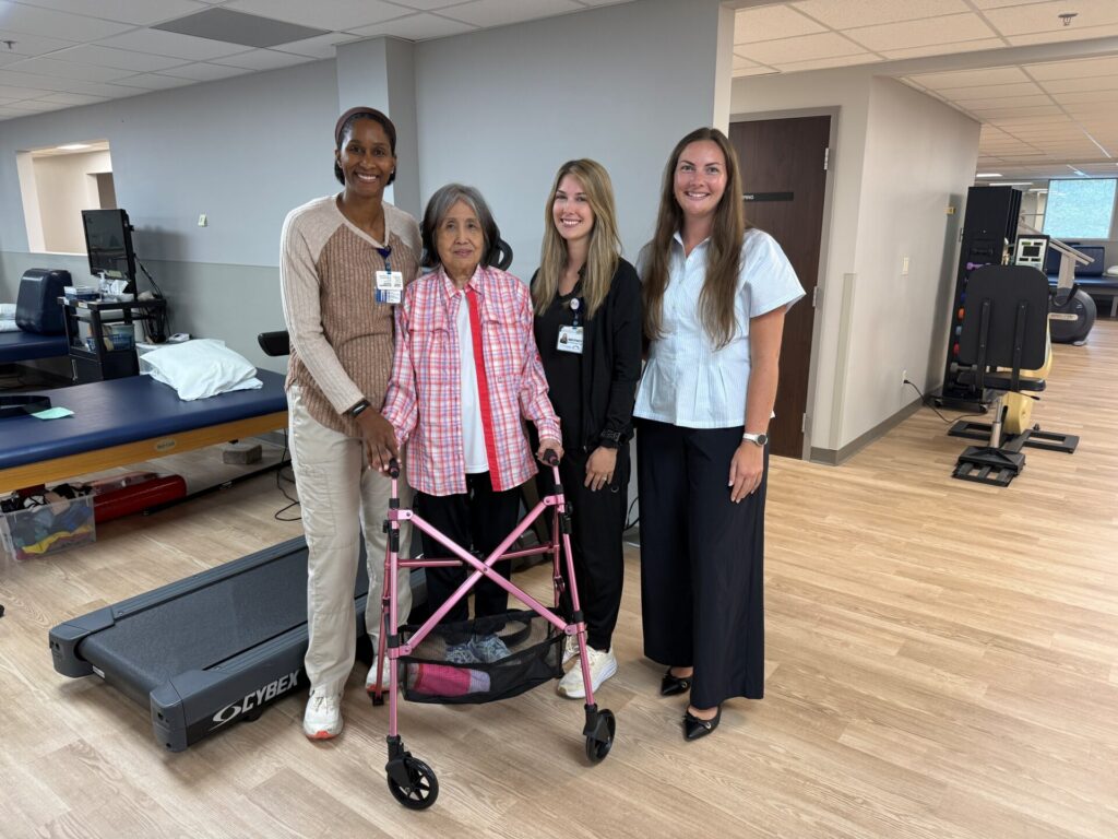 Four women stand together in a physical therapy room, one using a pink walker. Three wear name badges and business attire, and all are smiling at the camera. Therapy equipment is visible in the background.