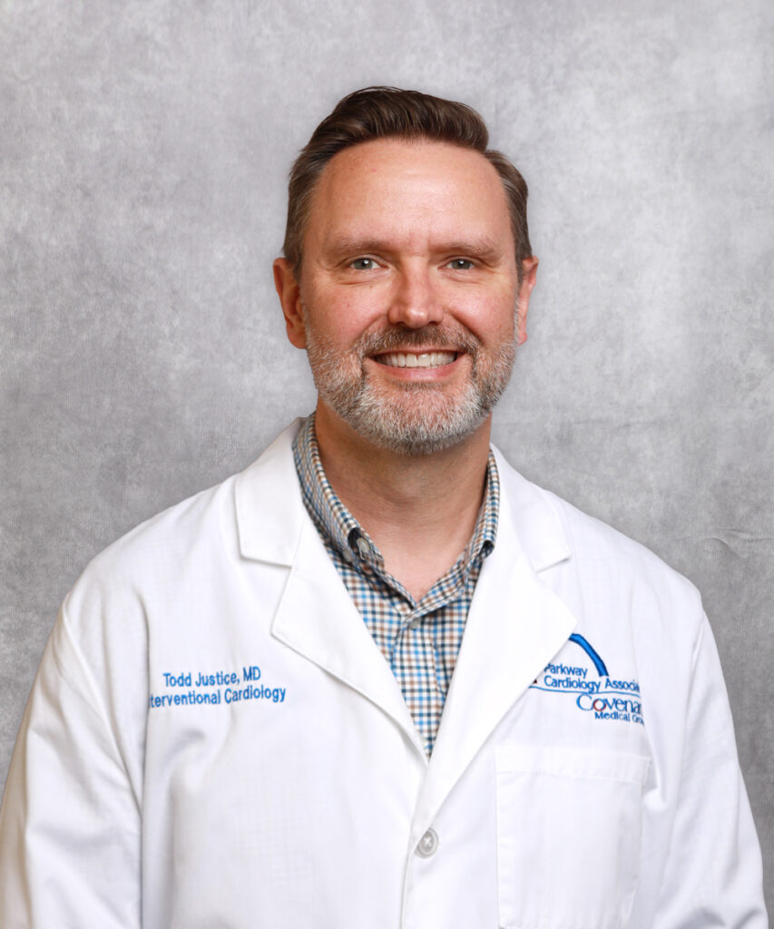 A smiling man with short brown hair and a beard wearing a white medical coat and plaid shirt stands in front of a light gray background.