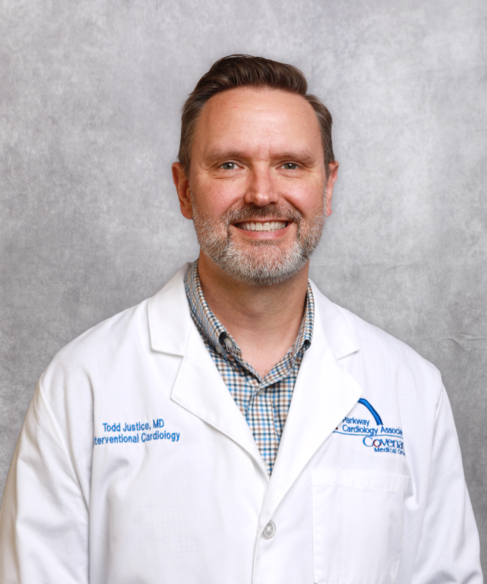 A smiling man with short brown hair and a beard wearing a white medical coat and plaid shirt stands in front of a light gray background.