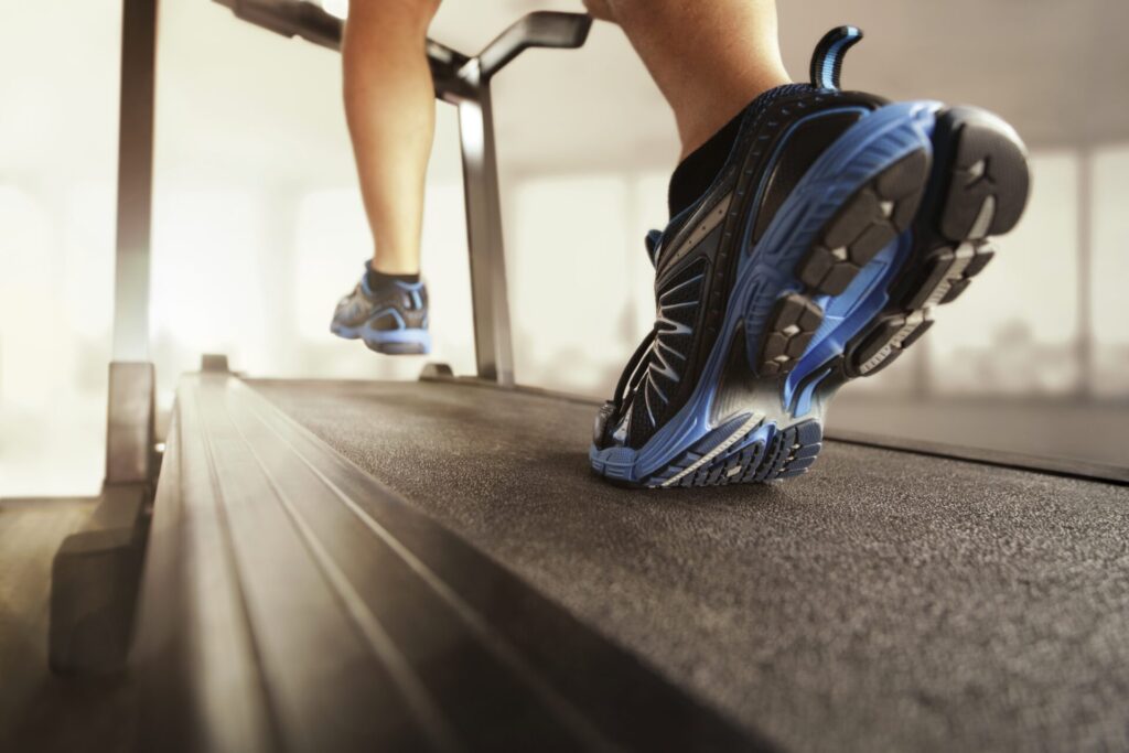 Close-up of a person’s legs and blue orthopedic running shoes as they walk or run on a treadmill in a gym, with a blurred background.