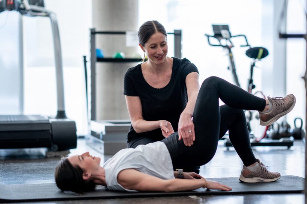 A woman lying on a mat does a bridge exercise while a trainer kneels beside her, offering guidance in a gym setting with equipment in the background.