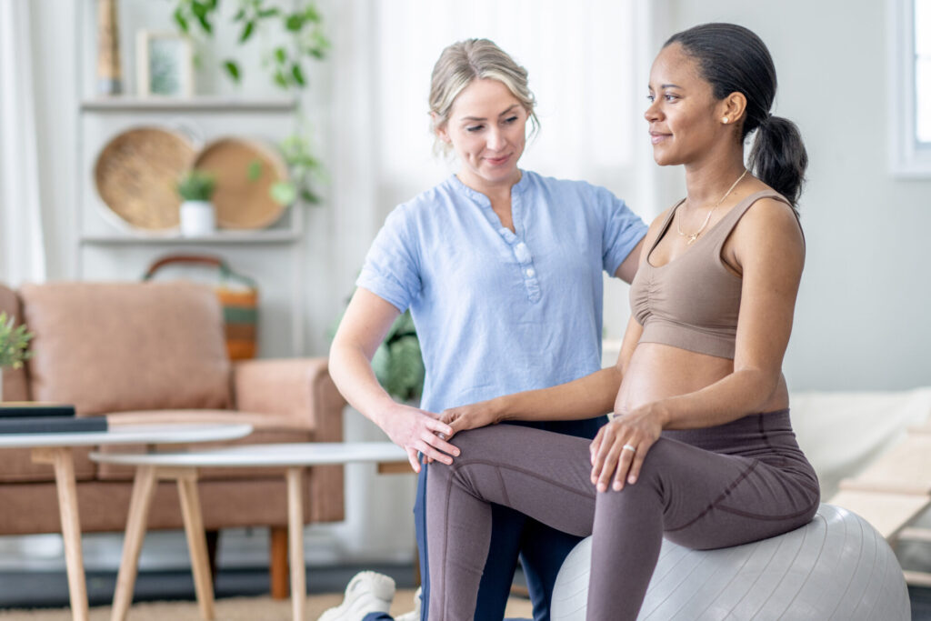 A pregnant woman sits on an exercise ball while a healthcare professional supports her knee and back, guiding her through a prenatal exercise in a bright, homey room.