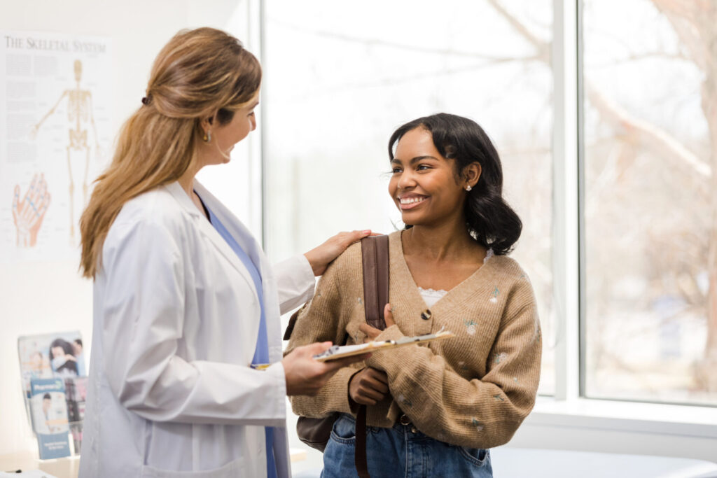 A doctor in a white coat smiles and talks with a young woman holding a bag. The doctor holds a clipboard. They stand in a bright medical office near a window.