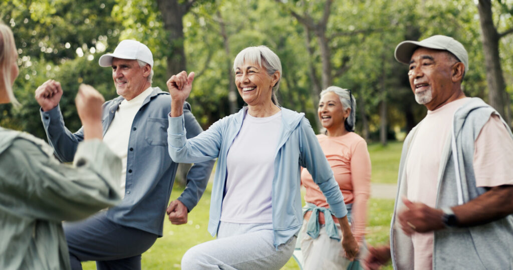 A group of smiling older adults exercises together outdoors in a park, lifting their knees and moving energetically—a testament to active living inspired by Advancing Hip Replacement at Covenant Health Morristown-Hamblen. The background is filled with green trees and grass.