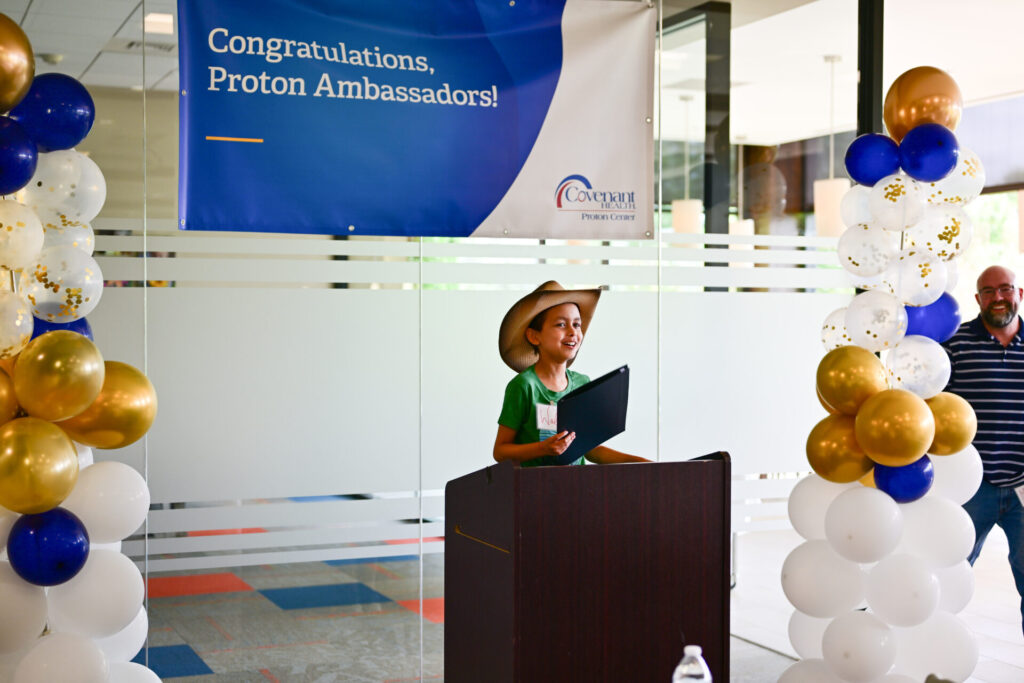 A person in a cowboy hat speaks at a podium under a banner reading Congratulations, Proton Ambassadors! with festive balloon columns on either side. A man stands in the background.