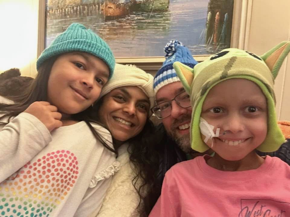 Four people, including two children in colorful hats, smile and pose closely for a selfie indoors. One child has a nasal tube and wears a green hat with Yoda ears. A painting hangs in the background.