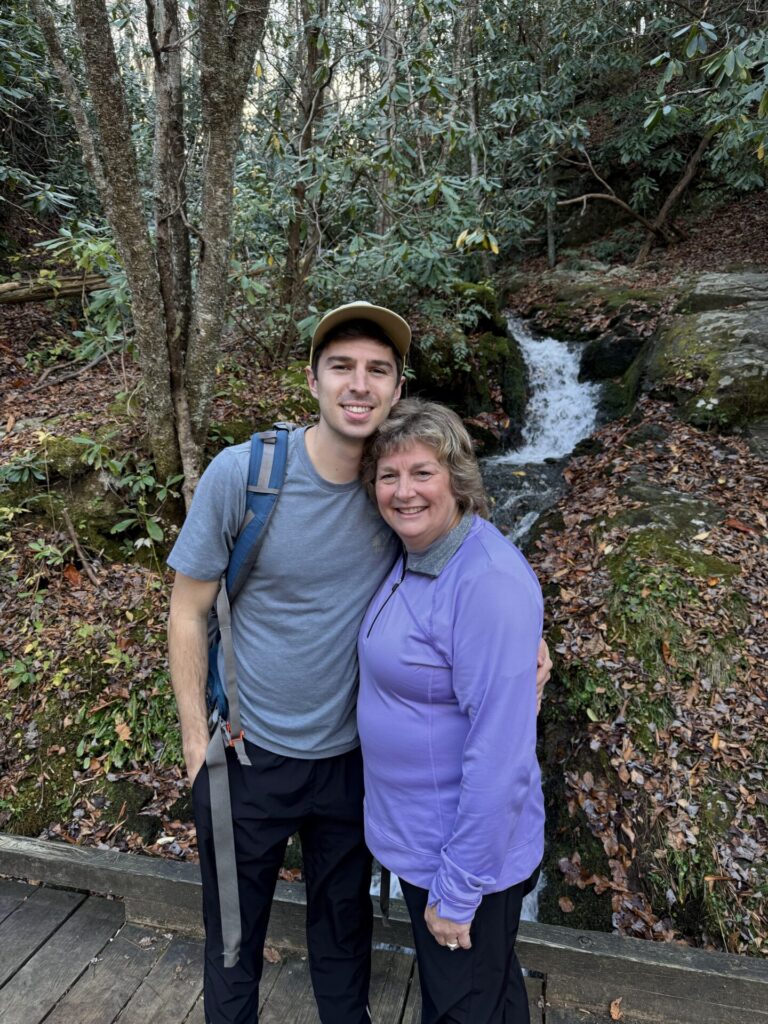 A young man and an older woman smiling and hugging in front of a small waterfall in a wooded area with fallen leaves on the ground.