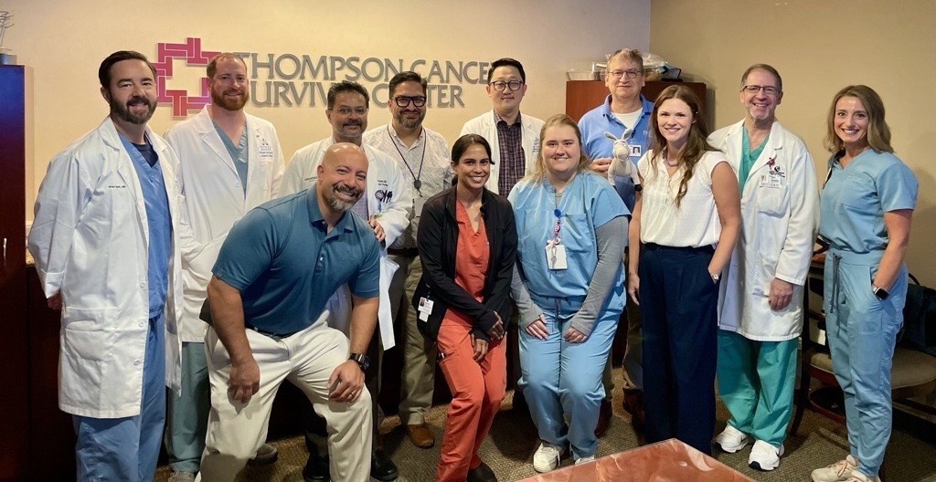 Group of 11 healthcare professionals from Fort Sanders Regional Medical Center and Thompson Cancer Survival Center pose together in an office beneath a Thompson Cancer Survival Center sign, marking their recent National Accreditation Program for Rectal Cancer (NAPRC) recertification. Team members wearing clinical scrubs, lab coats, and business attire stand and kneel for a group photo.