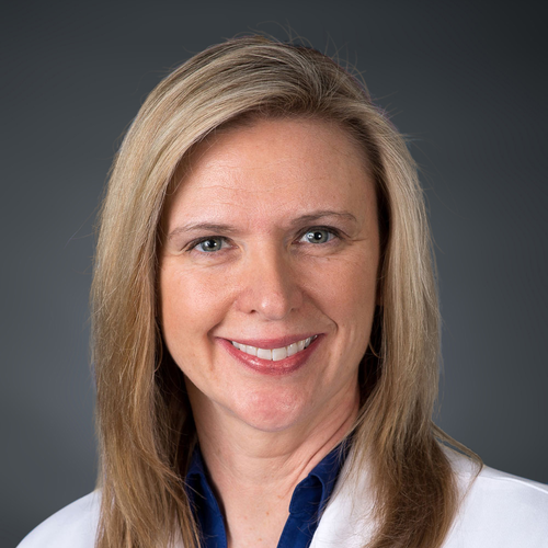 Smiling woman with straight, shoulder-length blonde hair, wearing a white coat and dark blue top, posed in front of a plain dark background.