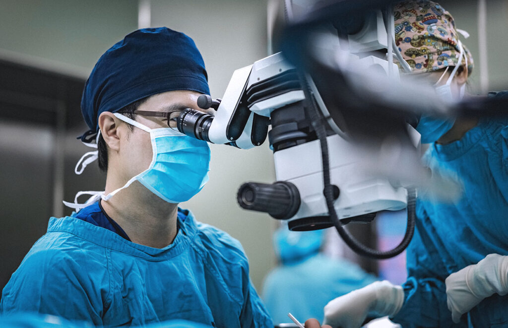 A surgeon wearing blue scrubs and a mask looks through a microscope, performing a medical procedure, while an assistant stands nearby.