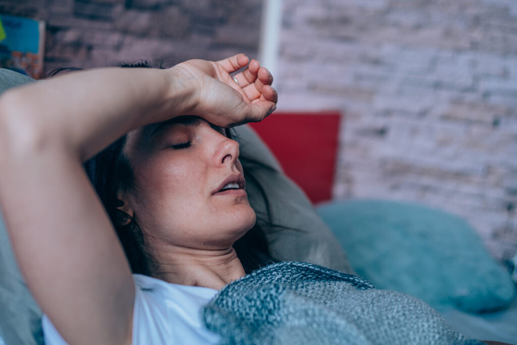 A woman lies on a couch with her eyes closed, covering her forehead with one hand, looking tired or unwell. Blankets and pillows are visible around her.