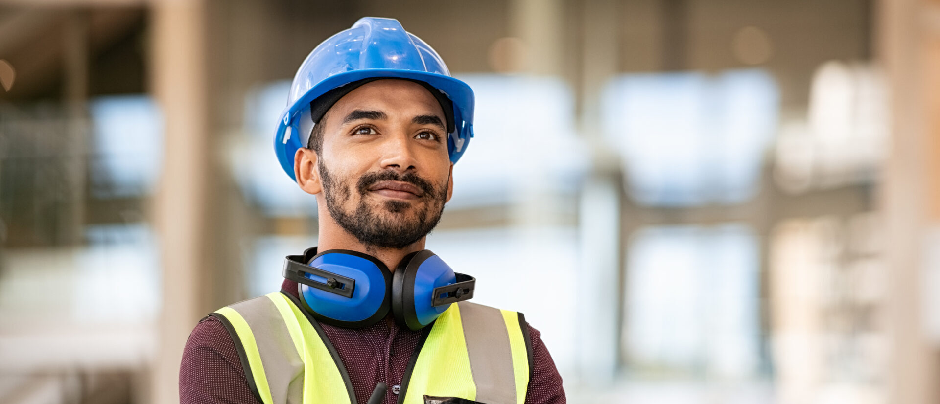 A man wearing a blue hard hat, safety vest, and headphones around his neck stands with arms crossed, looking confident. The background is blurred, suggesting an indoor construction or industrial setting.