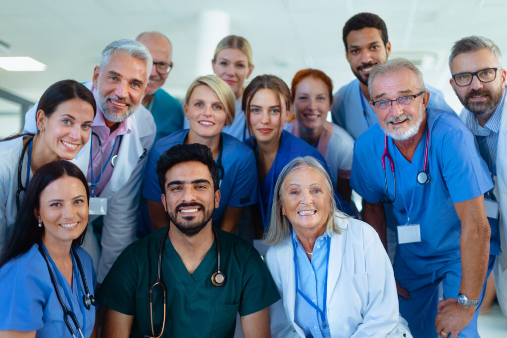 A diverse group of healthcare professionals, including doctors and nurses, smile together in a hospital setting, wearing scrubs and lab coats with stethoscopes and ID badges visible.