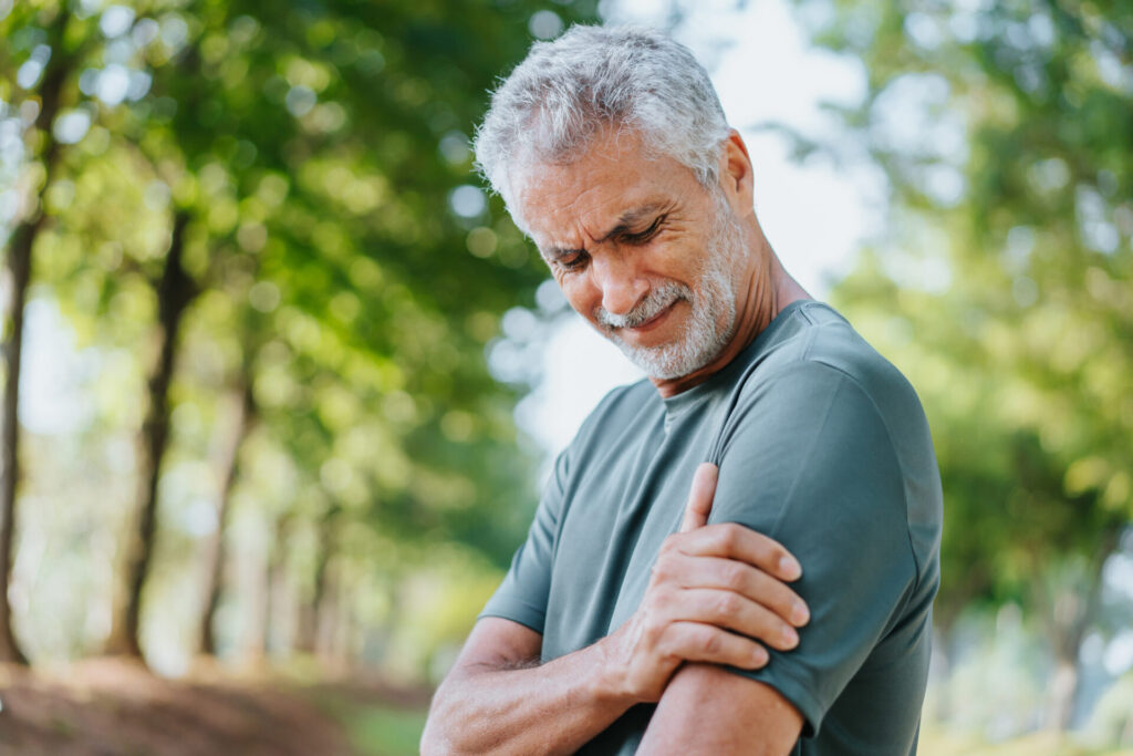 An older man stands outdoors, clutching his left arm with a pained expression, suggesting discomfort or pain. Trees and greenery are visible in the background—a reminder that recovery takes time, and sepsis fast treatment saves lives.