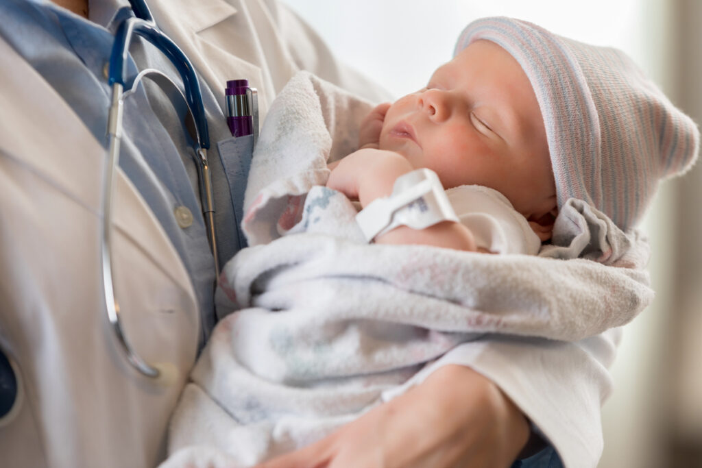 A newborn baby wearing a hospital hat and ID band is sleeping, wrapped in a blanket, being gently held by a medical professional with a stethoscope around their neck.