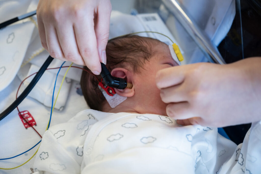 A newborn baby wearing a cloud-patterned onesie receives a hearing test; an adult is gently placing a device in the babys ear while the infant lies on a hospital bed with monitoring wires nearby.