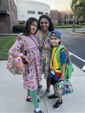 A woman standing outside with two smiling children, all wearing colorful outfits and backpacks, ready for school. The background shows a school building and greenery.
