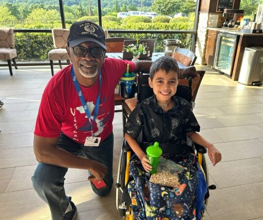 An older man kneels next to a smiling boy in a wheelchair. The boy holds a green cup and has a colorful blanket on his lap. They are indoors with large windows and greenery outside.